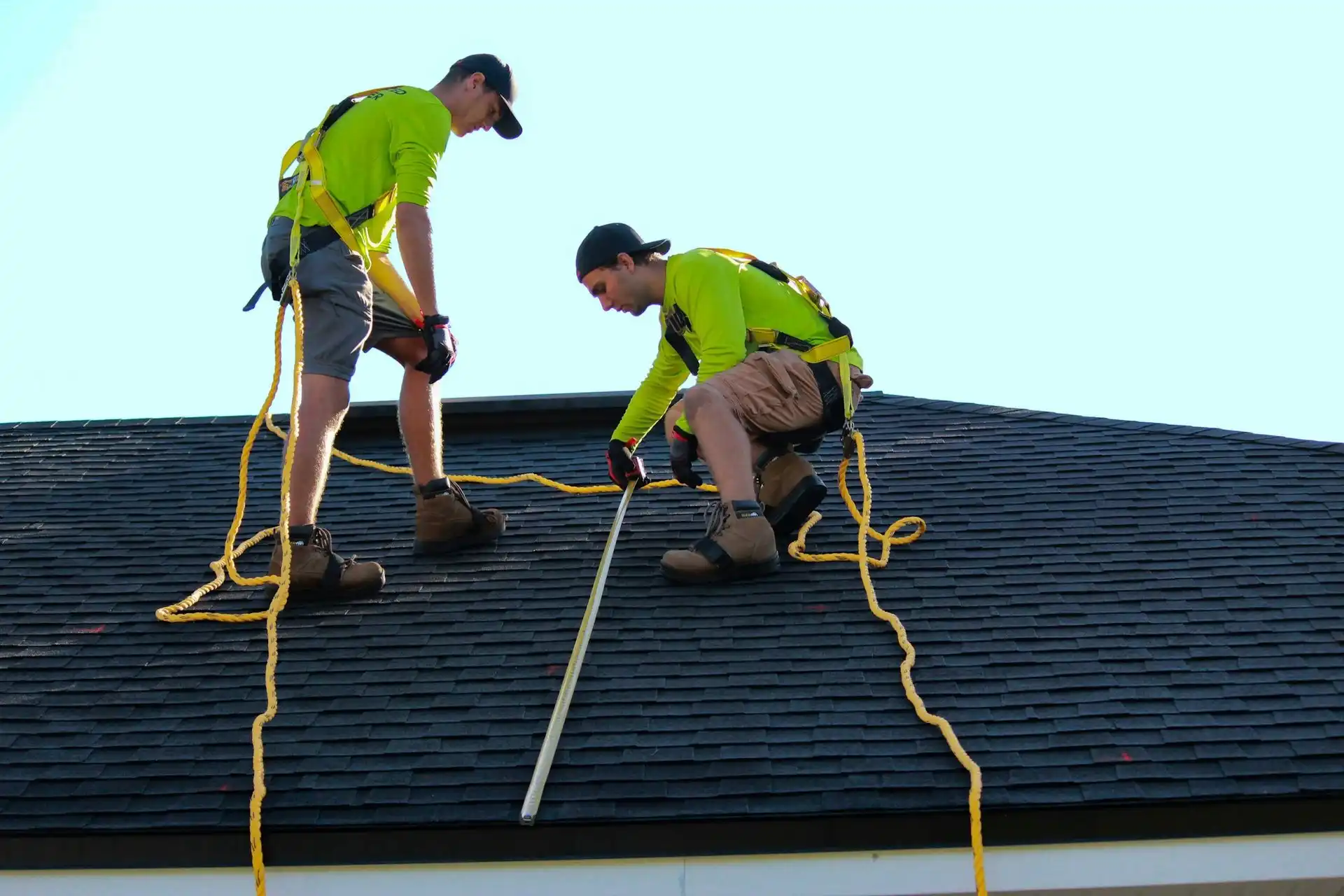 Two roofers in safety harnesses working on an asphalt shingle roof under a clear sky