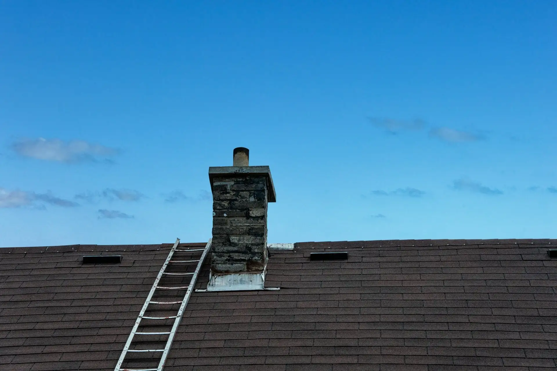 Ladder resting on a shingle roof next to a stone chimney, blue sky behind
