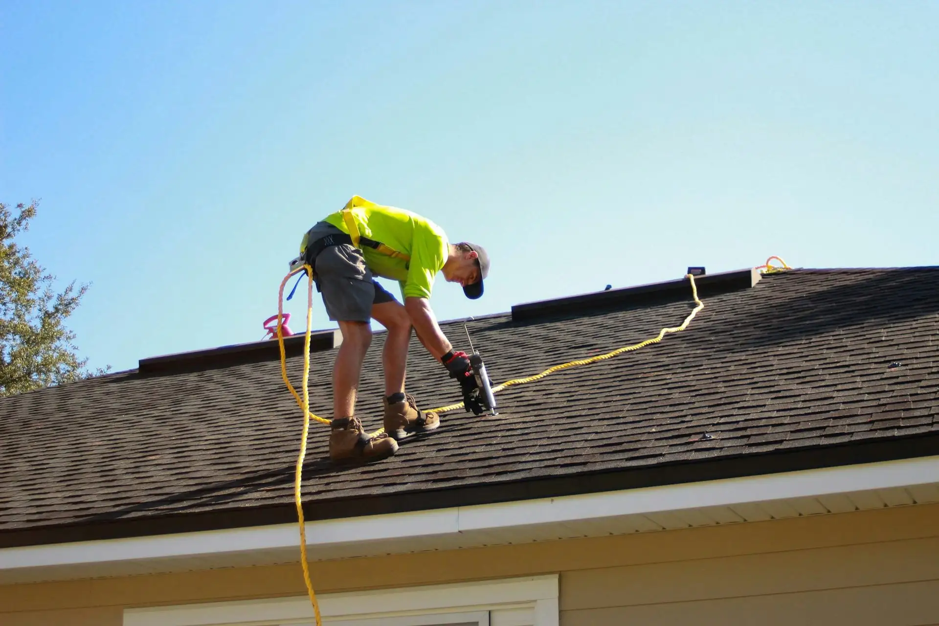 Roofer in a safety harness sealing a seam on a shingle roof with a caulk gun